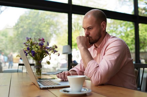 Thoughtful businessman work on notebook while sitting at wooden table in modern coffee shop interior, student reading text or book in cafe, male freelancer connecting to wireless via laptop computer