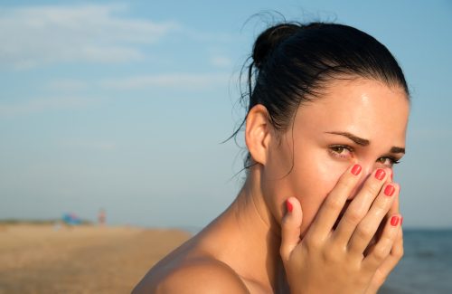 Close-up of a young woman with red eye  rubbing  irritated sensitive eyes or nose on the beach, allergy reaction